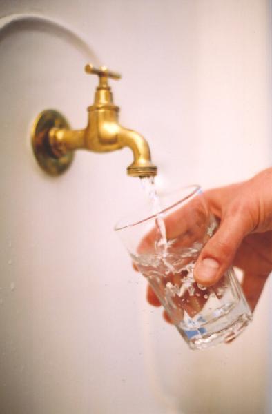 Eine Hand ist abgebildet, die ein Glas zu einem goldenen Wasserhahn hält. Aus dem Wasserhahn fließt Wasser in das Glas. 