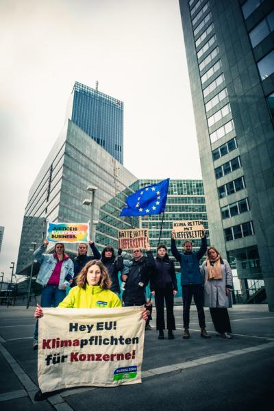 Anna Leitner steht im Vordergrund und hält ein Banner in die Kamera, darauf steht: "Hey EU! Klimapflichten für Konzerne!" Im Hintergrund stehen Menschen mit Fahnen und Banner