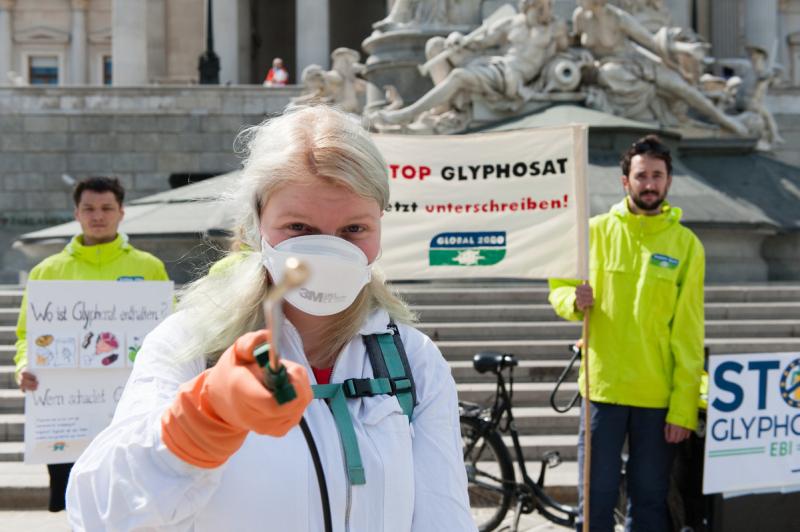 Frau mit Maske und Schutzanzug hält Pestizidsprühgerät in die Kamera. 