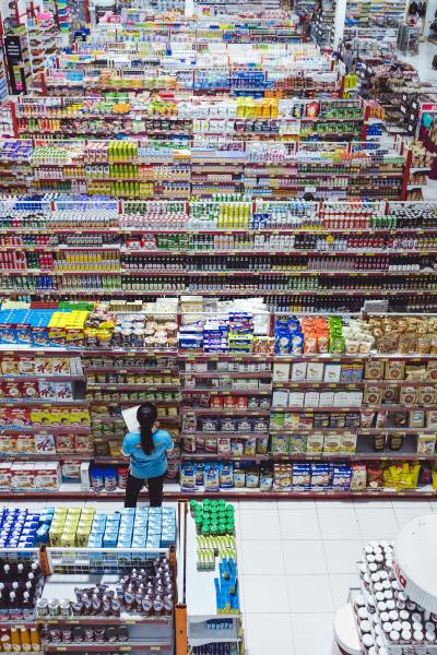 Totale von einem Supermarkt von Innen. Zahlreiche befüllte Regale hintereinander. Vorne steht eine Mitarbeiterin in einem blaue Tisht und schlichtet nach. 
