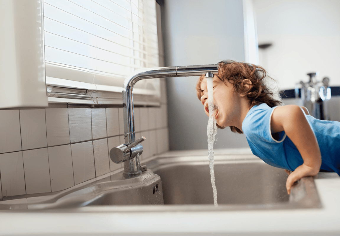 Kleines Kind mit braunen Haaren und blauem T-Shirt beugt sich über die Küchenzeile um aus dem Wasserhahn zu trinken. 