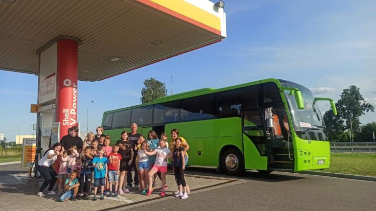 Kindergruppe mit Begleiter:innen stehen auf einer Tankstelle, im Hintergrund ein Reisebus