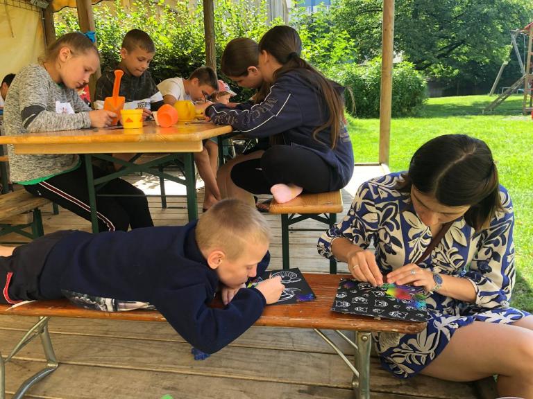 Kinder sitzen an einer Holzbank im Grünen und zeichnen