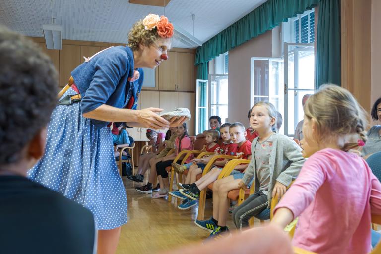 Eine Frau als Clown verkleidet bückt sich zu den Kindern herunter, die in einem Sesselkreis vor ihr sitzen