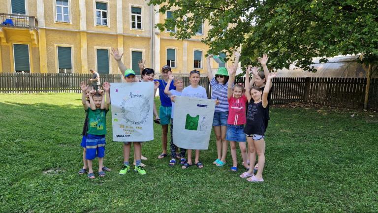 Kinder stehen in der Wiese vor einem Gebäude, sie präsentieren ihre Zeichnungen der Kamera