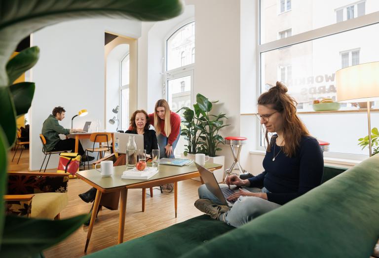 Eine Frau im Vordergrund sitzt auf dem Sofa und arbeitet am Laptop, der auf ihrem Schoß liegt. Im Hintergrund sieht man drei weitere Personen, die in der Arbeit vertieft sind. 