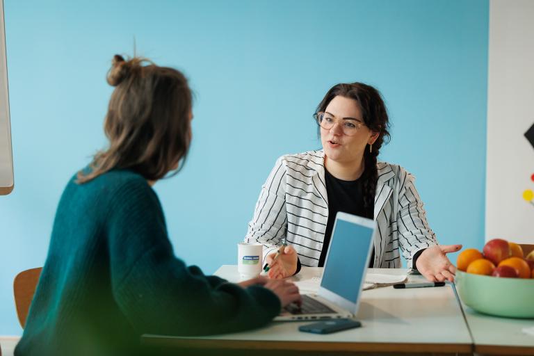 Zwei Frauen sitzen an einem Tisch und reden mit einander. Vor ihnen steht ein geöffneter Laptop 