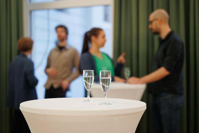 Im Fokus sind zwei Sektgläser auf einem Stehtisch mit Hussen. Im Hintergrund sieht man, wie sich zwei Paare unterhalten.