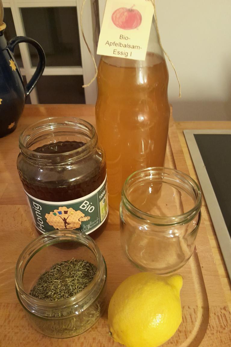 Auf einer Küchenzeile stehen eine Flasche Essig, ein Glas Honig, ein leeres Glas, ein Glas mit getrockneten Kräutern und eine Zitrone. 