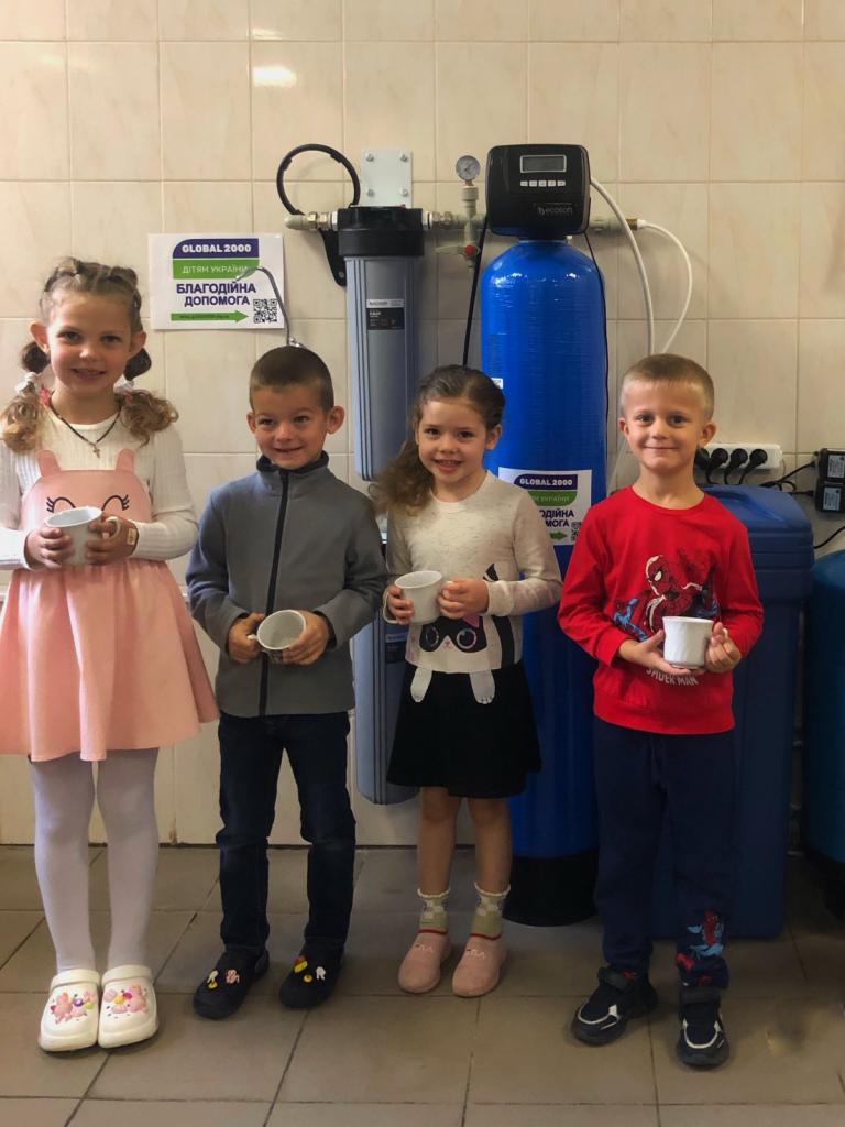 Vier Kinder stehen vor einer Wasseraufbereitungsanlage mit einem Glas Wasser in der Hand