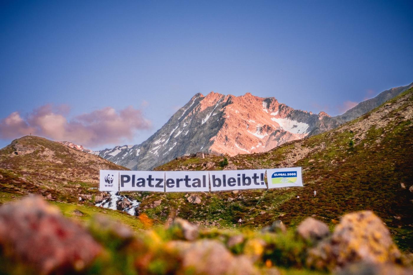 Das Platzertal, mit saftigen Wiesen und Blumen leuchtet in der Abendsonne. Ein riesiges Banner ist über das Tal gespannt, darauf befinden sich die Logos von WWF und GLOBAL 2000 und der Satz "Platzertal bleibt!". Im Hintergrund sieht man einen Berggipfel und der Himmel ist strahlend blau.