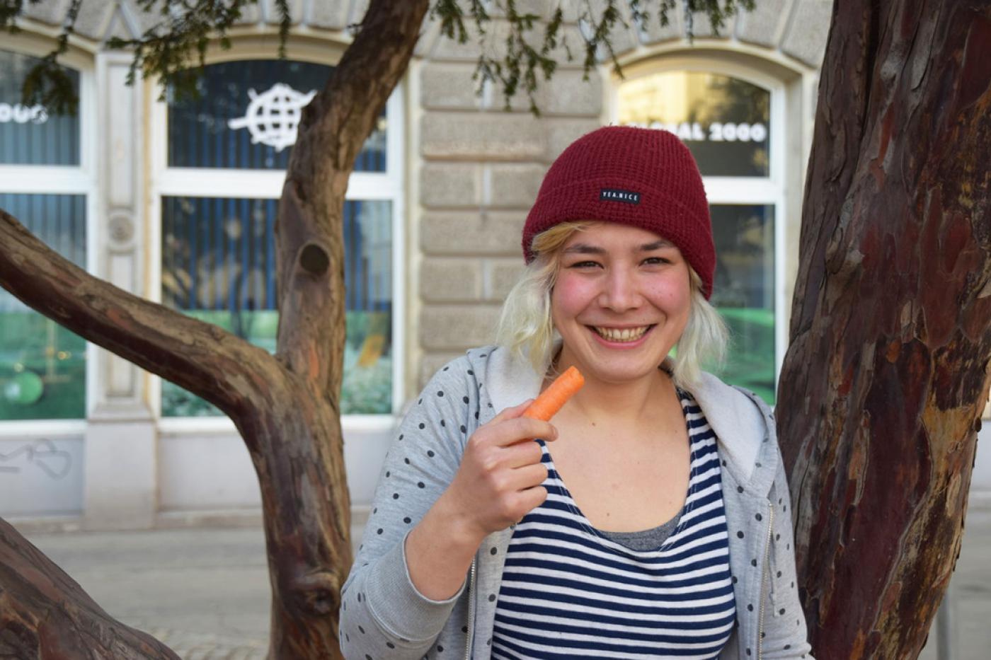 Blonde Junge Frau mit roter Haube und gestreiften Shirt sitzt auf einem Baum vor dem GLOBAL 2000 Gebäude und isst eine Karotte.