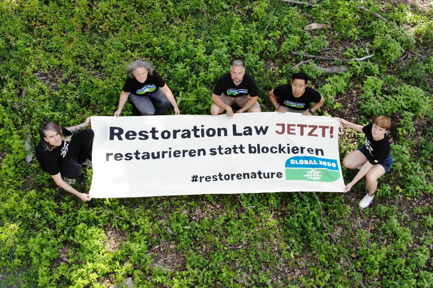 Foto aus der Vogelperspektive: 5 Aktivist*innen in GLOBAL 2000-T-Shirts halten einen großen Banner. Darauf steht "Restoration Law JETZT - restaurieren statt blockieren". 
