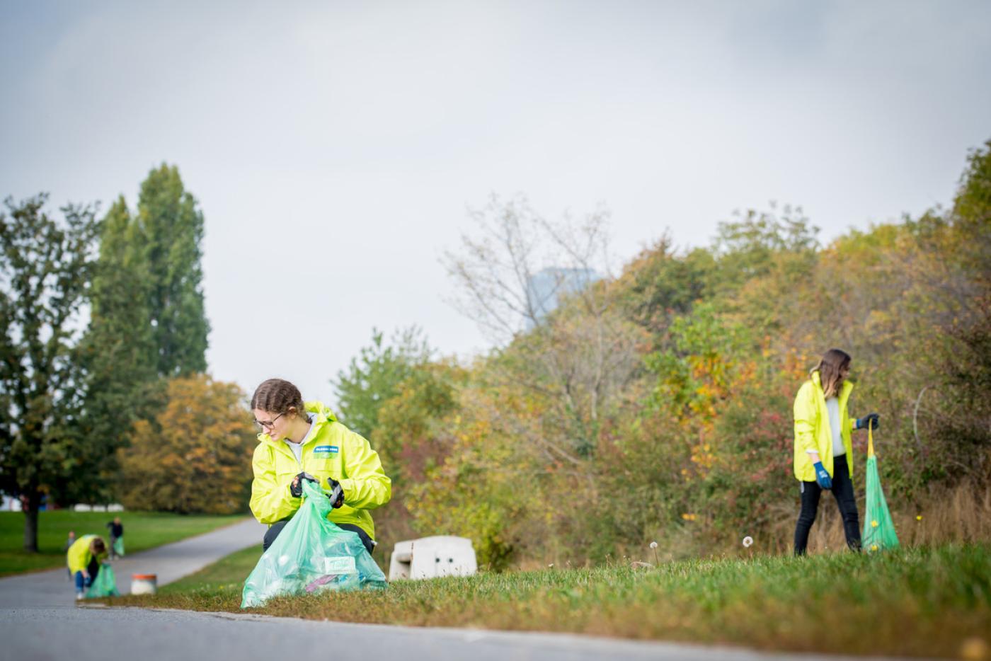 Drei Aktivit:innen mit gelben GLOBAL 2000 Jacken sammeln auf der Donauinel Müll
