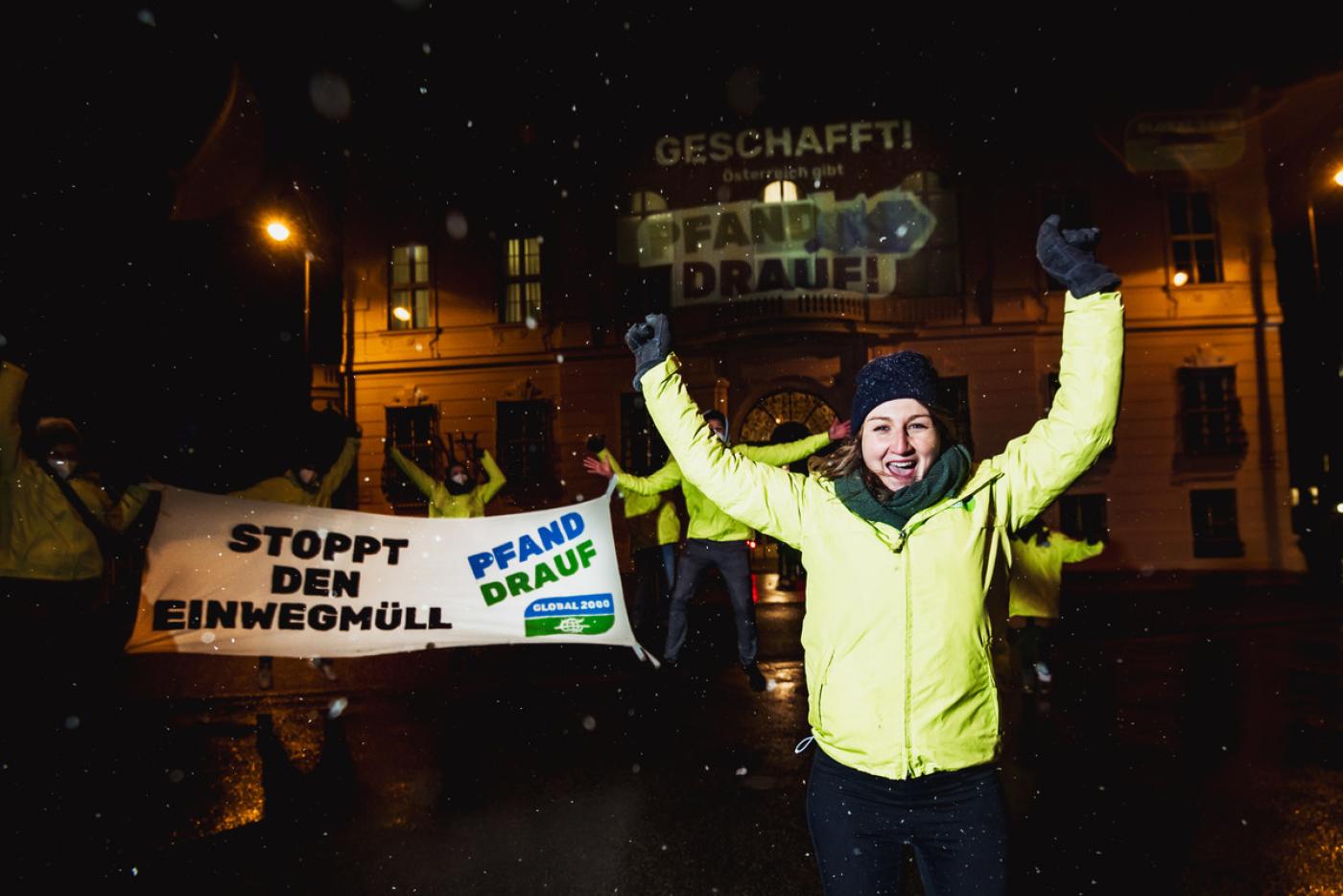 Eine Gruppe GLOBAL 2000 Aktivist:innen in gelben Jacken stehen vor dem Bundeskanzleramt in der Nacht und jubeln. Ein Banner auf dem "Stoppt den Einwegmüll" steht, wird von den Aktivist:innen gehalten. 