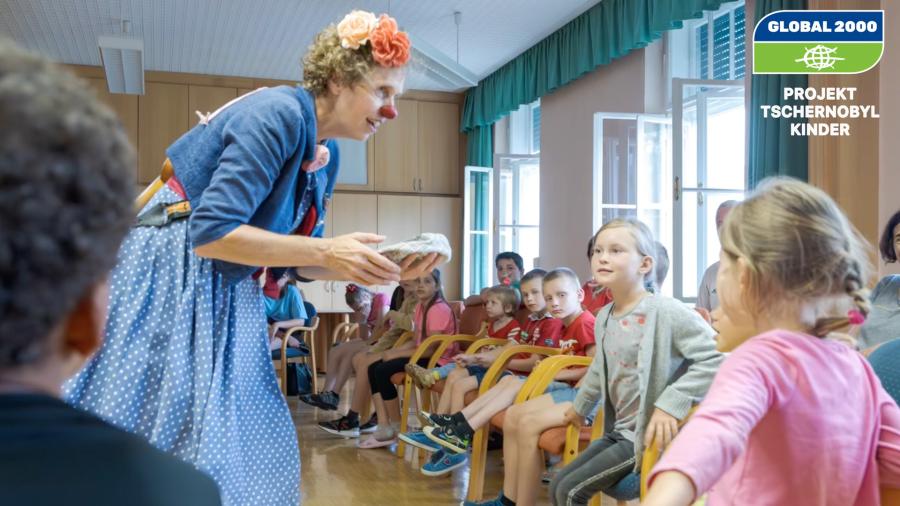 Kinder sitzen im Sesselkreis und schauen auf eine Frau die als Clown verkleidet ist. 