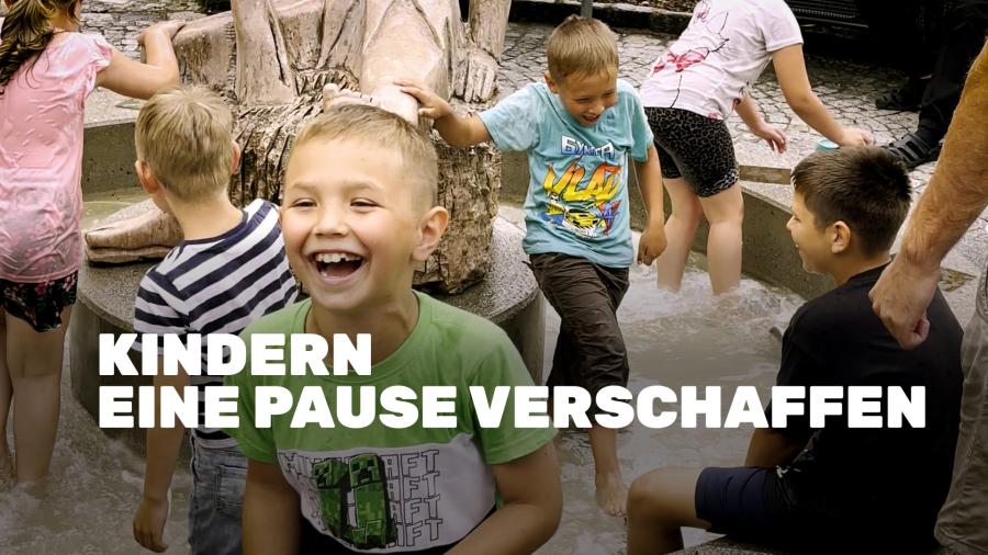 Ein junge mit grünem T-Shirt lacht ausgelassen in die Kamera. Im Hintergrund sieht man Kinder am Springbrunnen spielen. Darüber ist der Text: "Kindern eine Pause verschaffen"