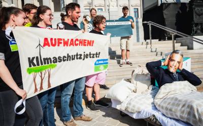 Aktivist:innen halten Banner in der Hand, worauf steht: "Aufwachen: Klimaschutz jetzt!". Ein Aktivist mit einer Nehammer-Maske liegt daneben im Bett und hält sich die Ohren zu.