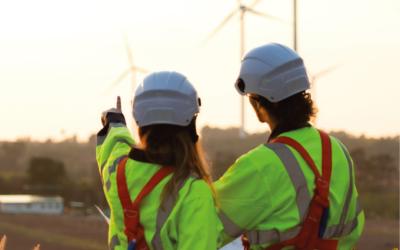 Zwei Ingeneur:innen in Arbeitskleidung schauen auf Windräder in der Ferne, sie sind mit dem Rücken zu uns gedreht und die Frau auf der linken Seite zeigt auf ein Windrad.