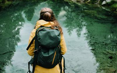 Frau mit gelber Regenjacke und Rucksack steht mit dem Rücken zur Kamera und schaut auf einen See, dahinter befinden sich die Berge im Nebel