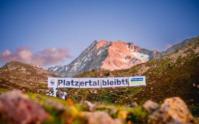 Das Platzertal, mit saftigen Wiesen und Blumen leuchtet in der Abendsonne. Ein riesiges Banner ist über das Tal gespannt, darauf befinden sich die Logos von WWF und GLOBAL 2000 und der Satz "Platzertal bleibt!". Im Hintergrund sieht man einen Berggipfel und der Himmel ist strahlend blau.