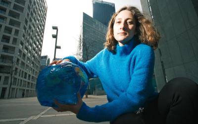 Anna Leitner trägt einen blauen Pullover, sie kniet am Boden vor hohen Bürogebäude und hält eine Plastik-Weltkugel in der Hand