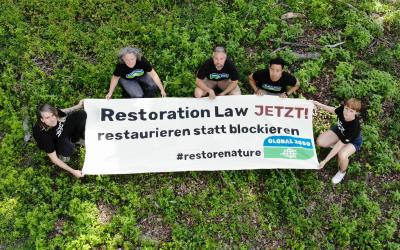 Foto aus der Vogelperspektive: 5 Aktivist*innen in GLOBAL 2000-T-Shirts halten einen großen Banner. Darauf steht "Restoration Law JETZT - restaurieren statt blockieren". 