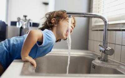 Kleines Kind mit braunen Haaren und blauem T-Shirt beugt sich über die Küchenzeile um aus dem Wasserhahn zu trinken. 