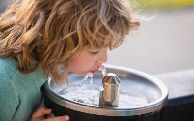 Ein blondes, lockiges Kind trinkt nach vorne gebeugt Wasser aus einem silbernen Wasserhahn an einem runden Waschbecken aus Edelstahl