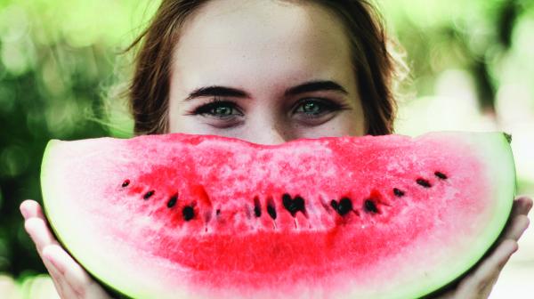 Eine braunhaarige Frau versteckt ihr Gesicht halb hinter einem Wassermelonenstück, das sie vor ihr gesicht hält.