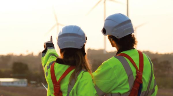 Zwei Ingeneur:innen in Arbeitskleidung schauen auf Windräder in der Ferne, sie sind mit dem Rücken zu uns gedreht und die Frau auf der linken Seite zeigt auf ein Windrad.