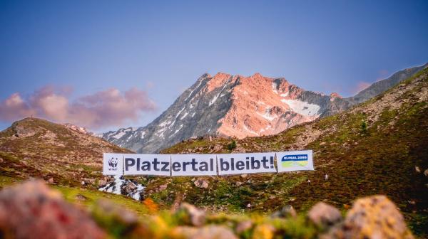 Das Platzertal, mit saftigen Wiesen und Blumen leuchtet in der Abendsonne. Ein riesiges Banner ist über das Tal gespannt, darauf befinden sich die Logos von WWF und GLOBAL 2000 und der Satz "Platzertal bleibt!". Im Hintergrund sieht man einen Berggipfel und der Himmel ist strahlend blau.