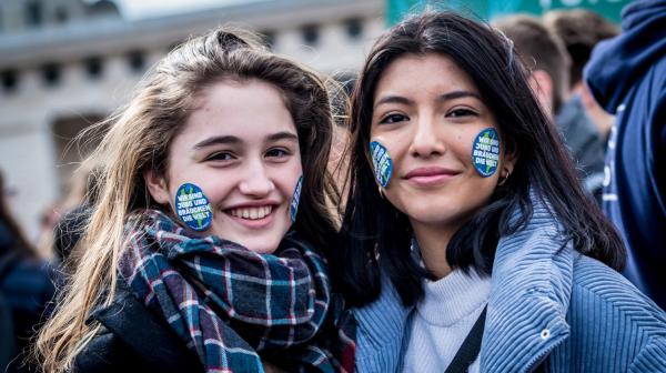 Zwei junge Frauen, stehen in der Menschenmenge einer Demo, sie sind warm angezogen und lächeln in die Kamera. Auf ihren Wangen kleben Sticker in Weltkugel-Optik.