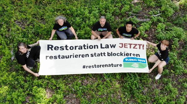 Foto aus der Vogelperspektive: 5 Aktivist*innen in GLOBAL 2000-T-Shirts halten einen großen Banner. Darauf steht "Restoration Law JETZT - restaurieren statt blockieren". 