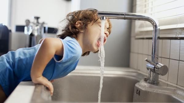 Kleines Kind mit braunen Haaren und blauem T-Shirt beugt sich über die Küchenzeile um aus dem Wasserhahn zu trinken. 