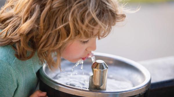 Ein blondes, lockiges Kind trinkt nach vorne gebeugt Wasser aus einem silbernen Wasserhahn an einem runden Waschbecken aus Edelstahl